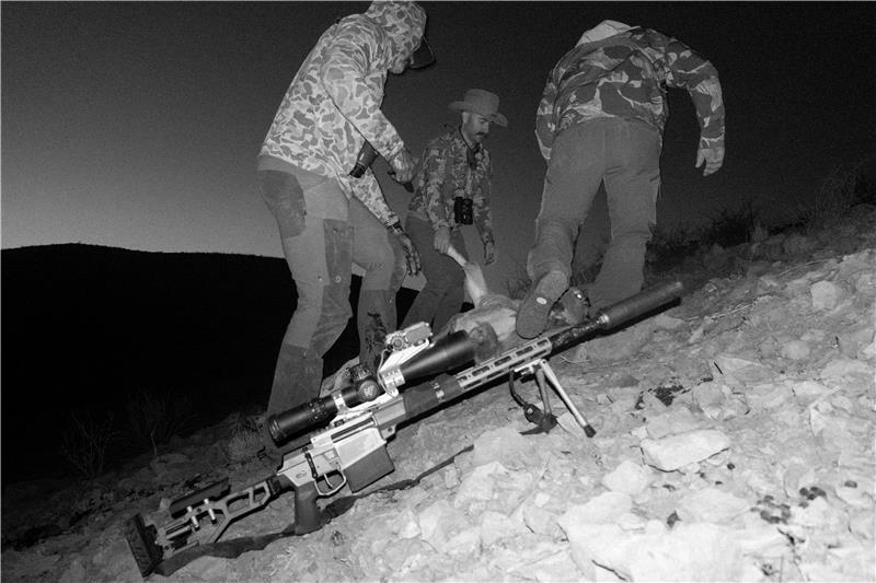 Three hunters work together on a rocky hillside at dusk to recover a harvested aoudad, while a precision rifle with a suppressor rests in the foreground.