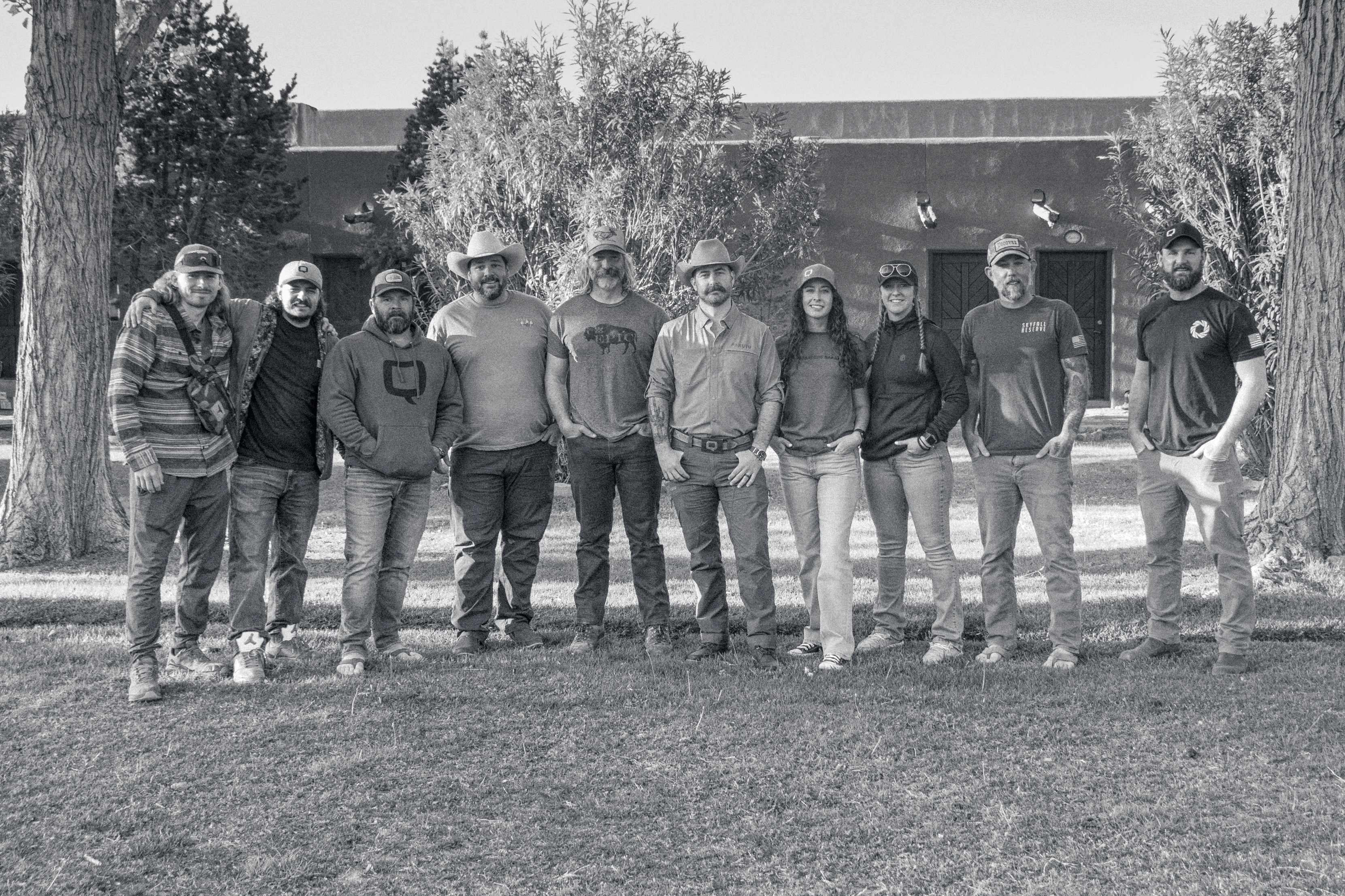 The Q team stands together in a line on a grassy lawn, wearing casual outdoor and branded apparel, with trees and a rustic building in the background. The black-and-white photo captures the team in a relaxed and unified pose.
