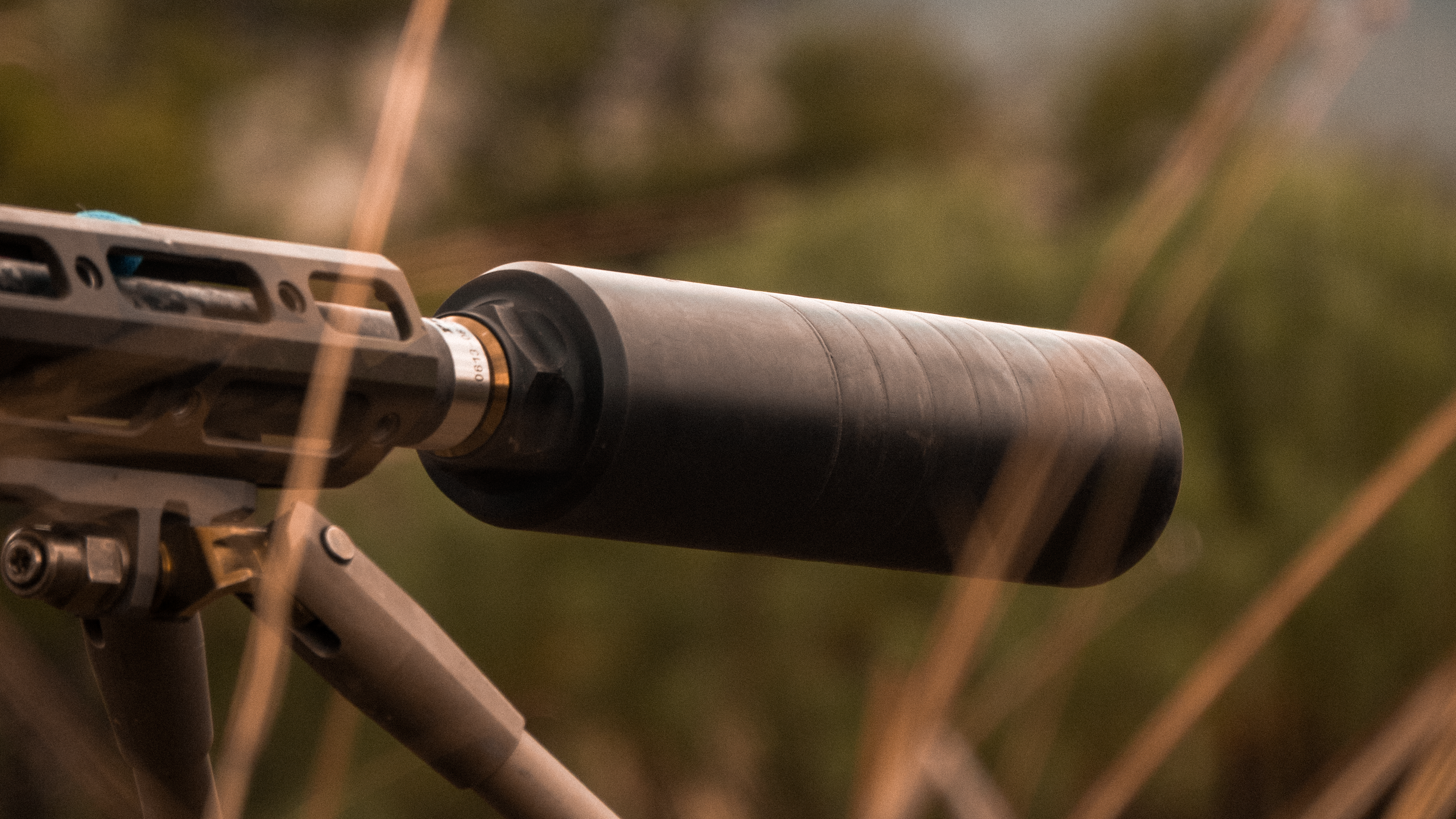Close-up shot of the Speakeasy suppressor mounted on a precision rifle in a rugged outdoor setting. The image focuses on the rear portion of the suppressor and its attachment point, showcasing its sturdy build and secure Quickie Fast-Attach system. Earth tones and dry grasses frame the shot, reinforcing the suppressor’s readiness for real-world field use.