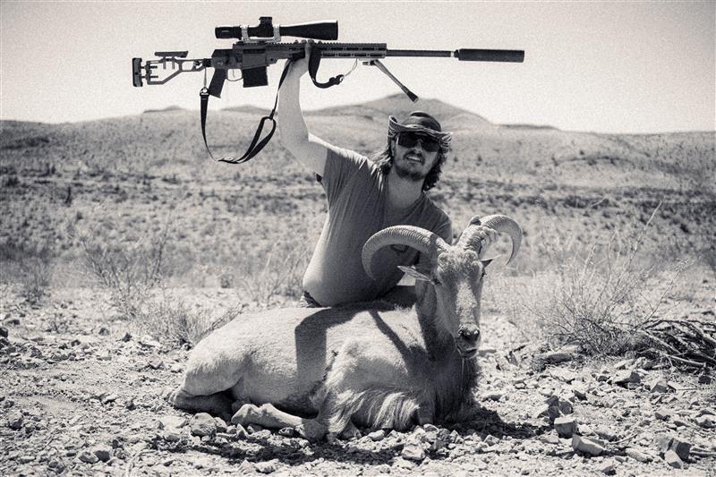 Matt kneels beside a harvested aoudad in the rocky terrain of West Texas, holding a suppressed precision rifle triumphantly overhead. The black-and-white photo captures the rugged spirit of the hunt.