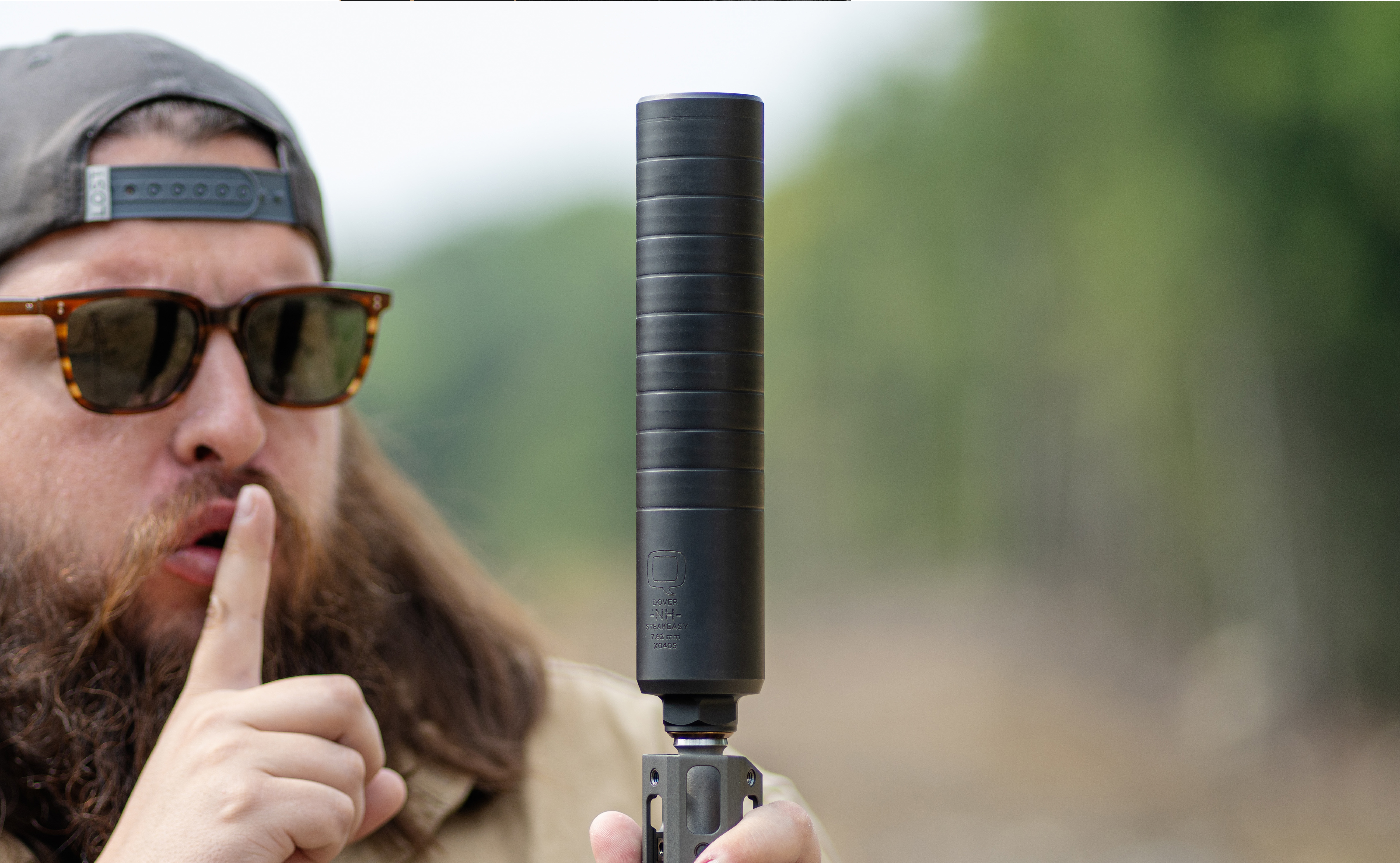 A man in sunglasses and a backwards cap holds a rifle equipped with the Speakeasy suppressor. With his index finger raised to his lips in a “shh” gesture, the image reinforces the product’s quiet performance. The background features a blurred forested shooting range, emphasizing the suppressor's outdoor utility and stealth.