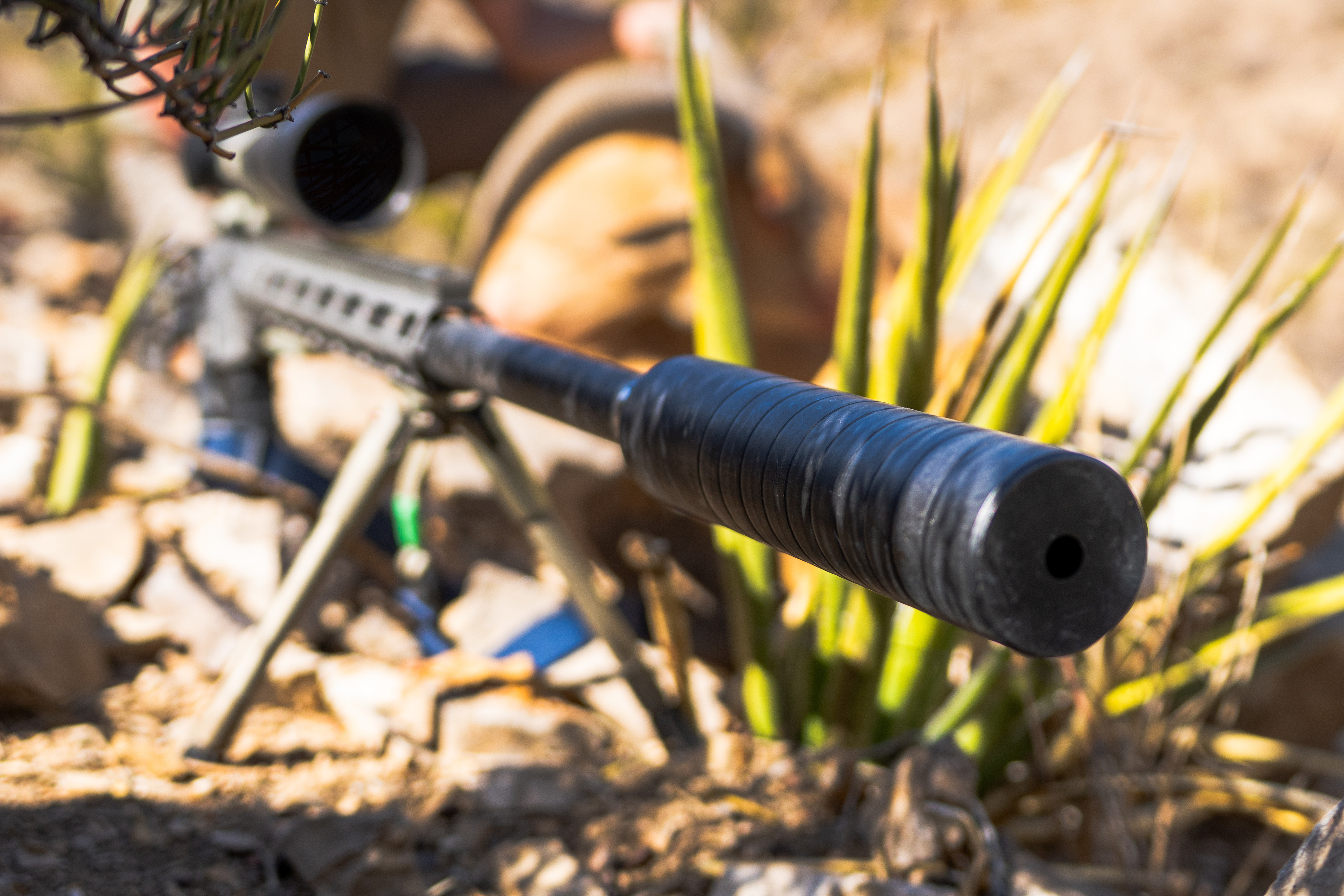 Close-up of a rifle equipped with the Speakeasy suppressor, captured in a desert environment. The image focuses on the suppressor from the front angle, showcasing its matte black finish and grooved exteri